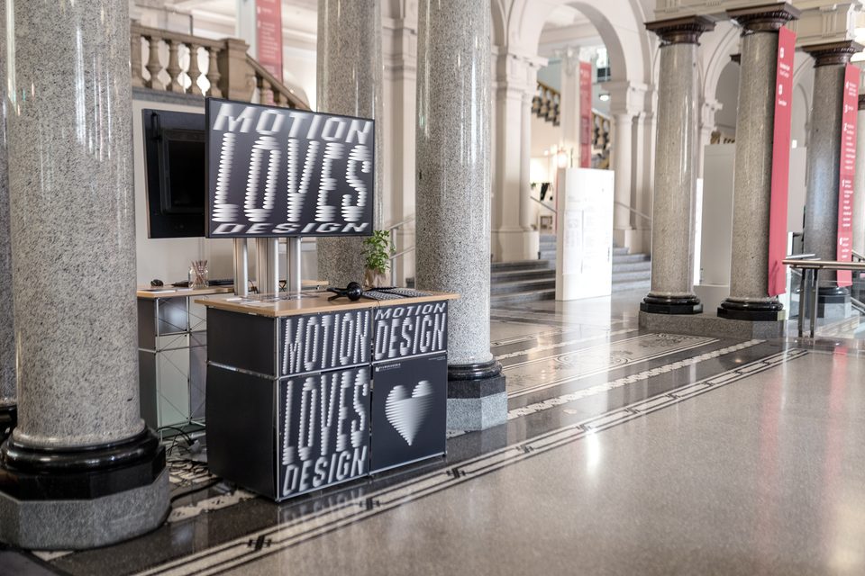 side view of the exhibition booth between columns, with a screen and labeled panels reading „Motion Loves Design“